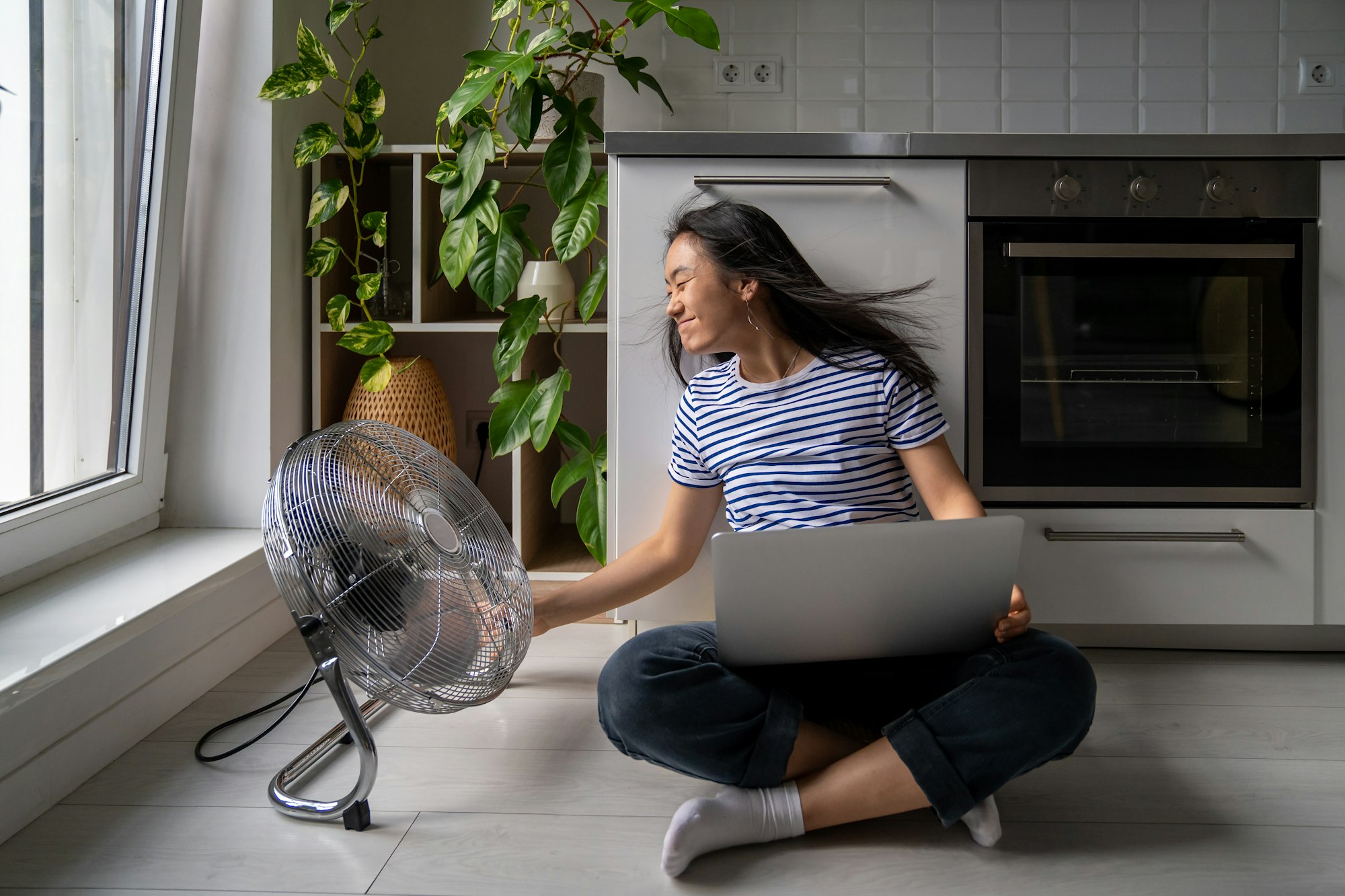 Girl turned powerful electric fan enjoying cool wind sitting on floor home work with laptop.